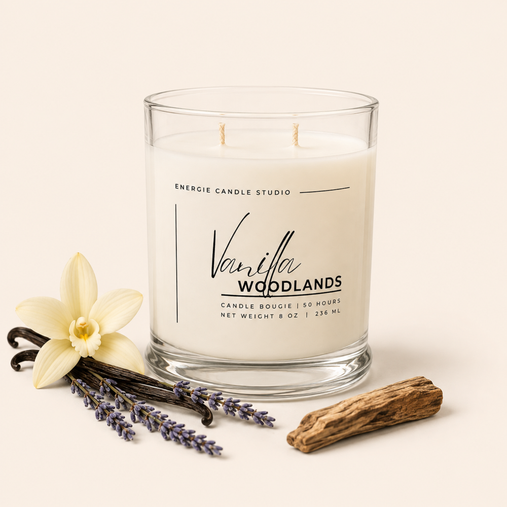 A two-wick scented candle in a clear glass jar by Energie Candle Studio, labeled Vanilla Woodlands. The candle is styled on a warm cream background with a vanilla flower, lavender sprigs, a vanilla bean, and a piece of driftwood.