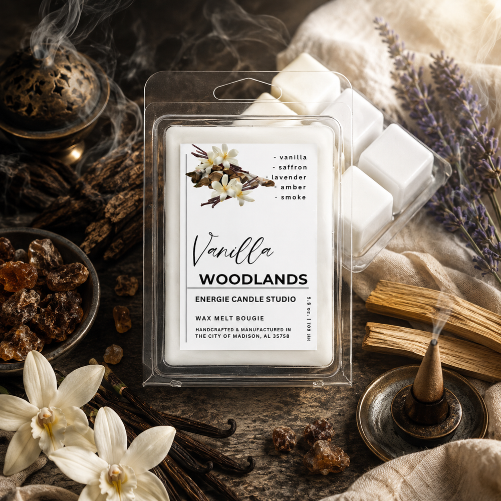 A wax melt package labeled 'Vanilla Woodlands' by Energie Candle Studio, shown in a clear clamshell with white wax cubes beside it. The scene is styled with dried flowers, incense, wood pieces, and soft fabric on a rustic surface.