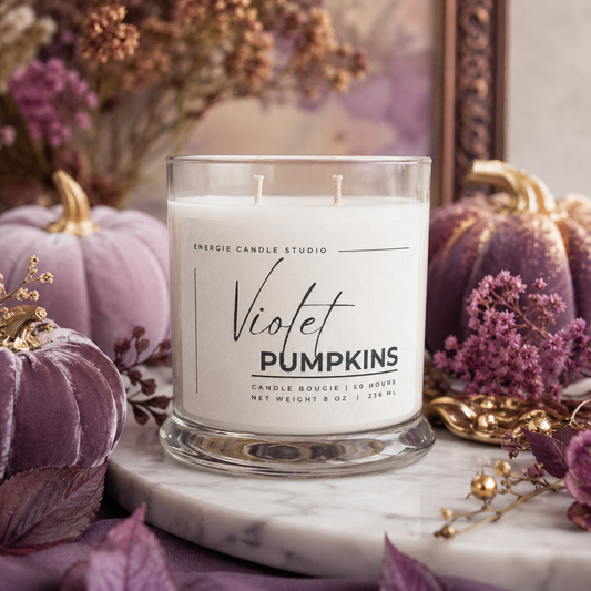 A double-wick candle in a clear glass jar labeled 'Violet Pumpkins' by Energie Candle Studio, styled on a marble surface with purple velvet pumpkins, dried flowers, and gold accents in a fall-themed setting.