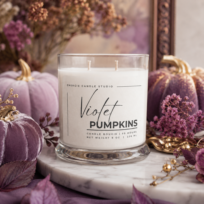 A double-wick candle in a clear glass jar labeled 'Violet Pumpkins' by Energie Candle Studio, styled on a marble surface with purple velvet pumpkins, dried flowers, and gold accents in a fall-themed setting.