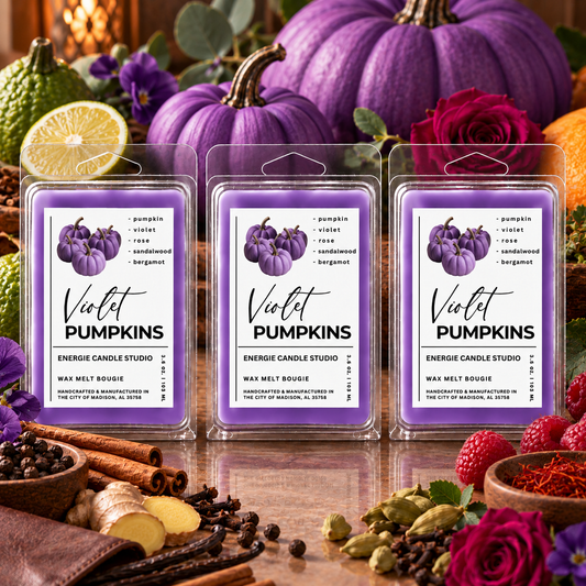 Three clear plastic clamshell packages of wax melts labeled 'Violet Pumpkins' are arranged on a reflective surface with autumn ingredients and large purple pumpkins in the background. The labels are purple with a pumpkin illustration and black text.