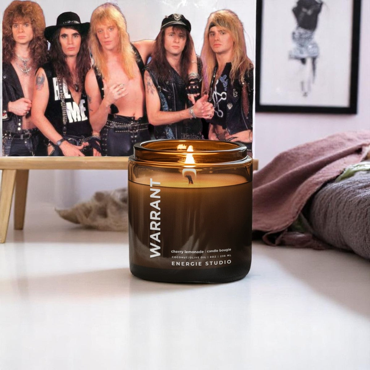 a glass candle  in a bedroom with with a picture of the band warrant on a wooden easel and a pink blanket in the background. 