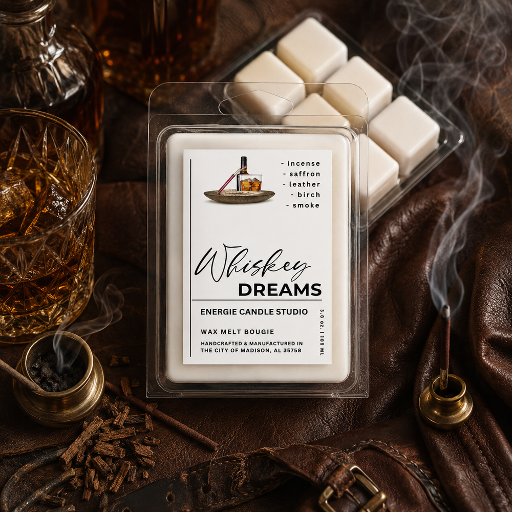 A packaged wax melt product labeled 'Whiskey Dreams' by Energie Candle Studio, placed on a dark brown leather surface with whiskey glassware, a brass incense holder, and white wax melts visible in a clear tray. The scene is styled with warm, moody lighting and smoke.