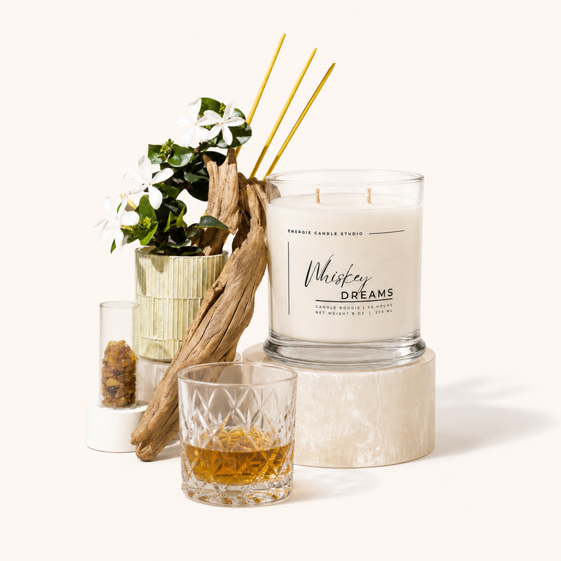 A large clear glass jar candle labeled 'Whiskey Dreams' by Smokey Candle Studio, styled on a light stone pedestal with a small whiskey glass, dried reeds, driftwood, and white flowers against a warm off-white background.