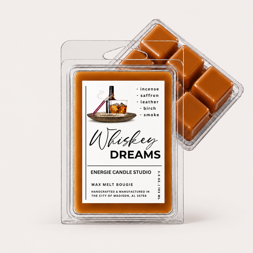 Two clear plastic clamshell wax melt packs on a light warm gray background. The front pack has a white label reading 'Whiskey Dreams' with a whiskey bottle illustration and scent notes including saffron, leather, birch, and smoke.
