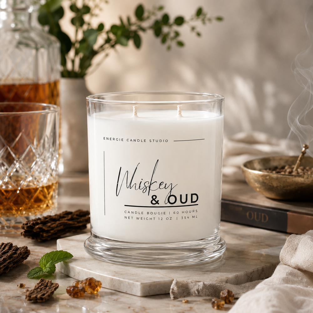 A white wax candle in a clear glass jar labeled 'Whiskey & Oud' by Energie Candle Studio, styled on a marble surface with whiskey glassware, dried herbs, mint, and a book in a warm indoor setting.