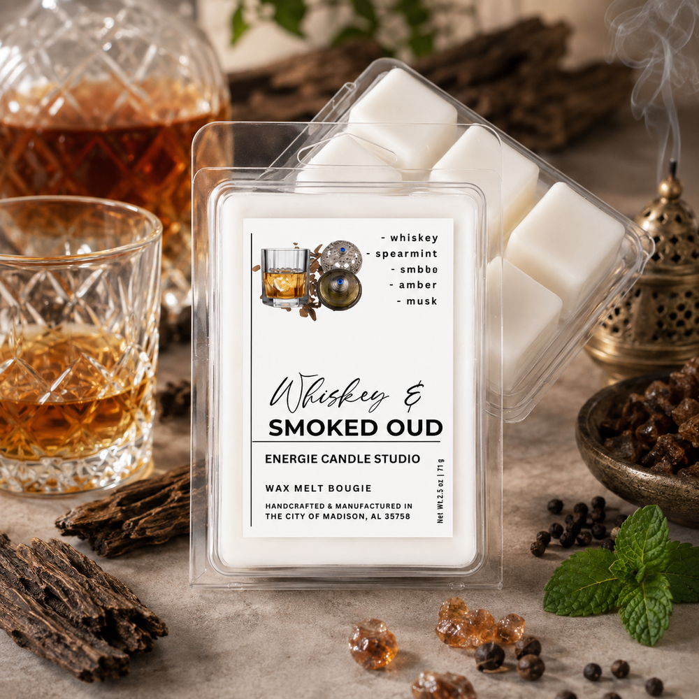 A clear plastic clamshell package of Energize Candle Studio 'Whiskey & Smoked Oud' wax melts sits on a stone surface with a second package behind it. The front label shows a whiskey glass illustration and scent notes: whiskey, spearmint, smoke, amber, and musk.